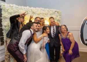 Bride and groom posing with family and friends in front of a white floral backdrop while taking a group photo at a wedding photo booth.